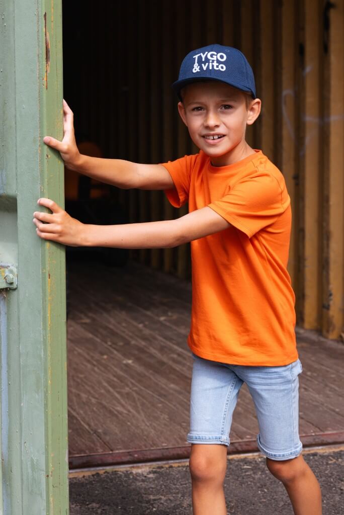 Boy wearing an orange shirt and blue cap with visible branding and light blue denim shorts Quinn, standing inside a wooden structure.