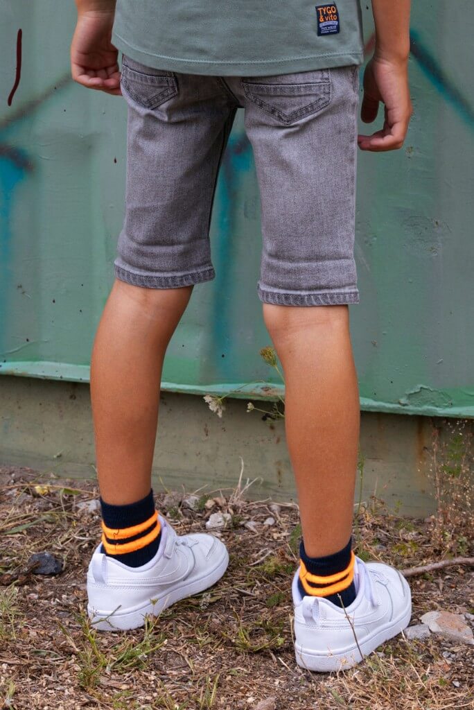 boy wearing gray shorts Quinn, white sneakers, and striped socks standing on a grassy area with a green wall in the background.