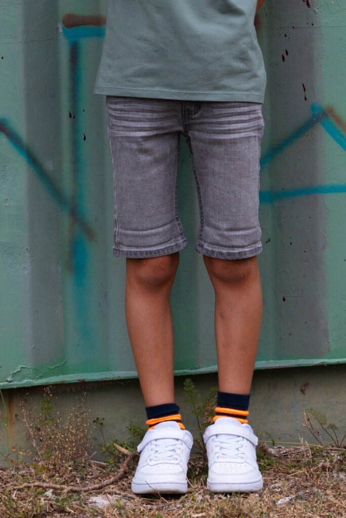 Boy wearing gray shorts Quinn and white sneakers standing against a textured wall with graffiti.