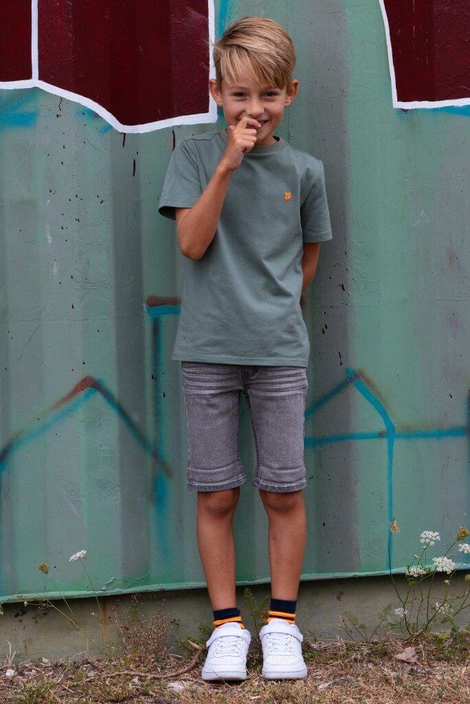Boy wearing a green t-shirt and gray shorts standing in front of a graffiti-covered wall.