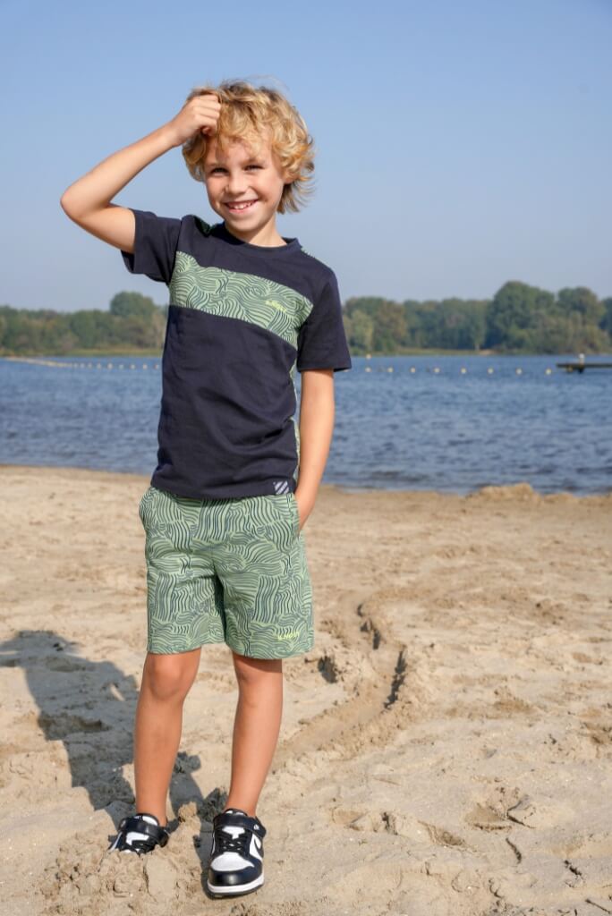 Boy wearing a navy t-shirt Tanner by B.Nosy with green design and patterned shorts standing on a sandy beach.