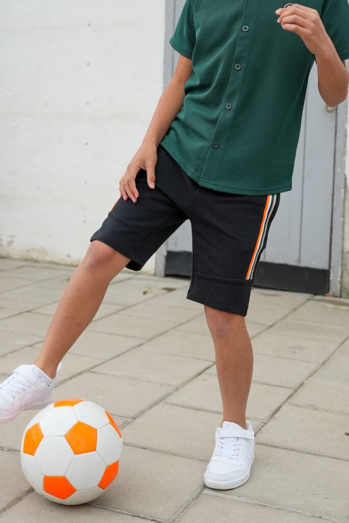 Boy wearing a green shirt and black shorts Bing with an orange stripe, standing next to a soccer ball on a paved surface.