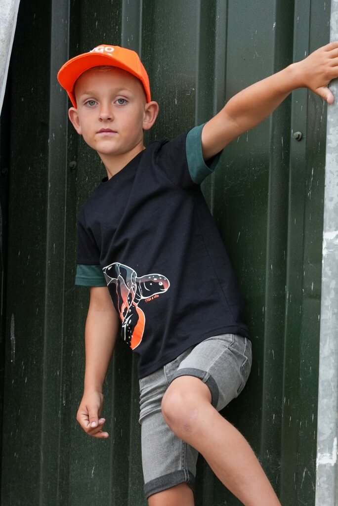 A boy wearing an orange cap and black t-shirt Toby with a graphic design, leaning against a dark green wall.
