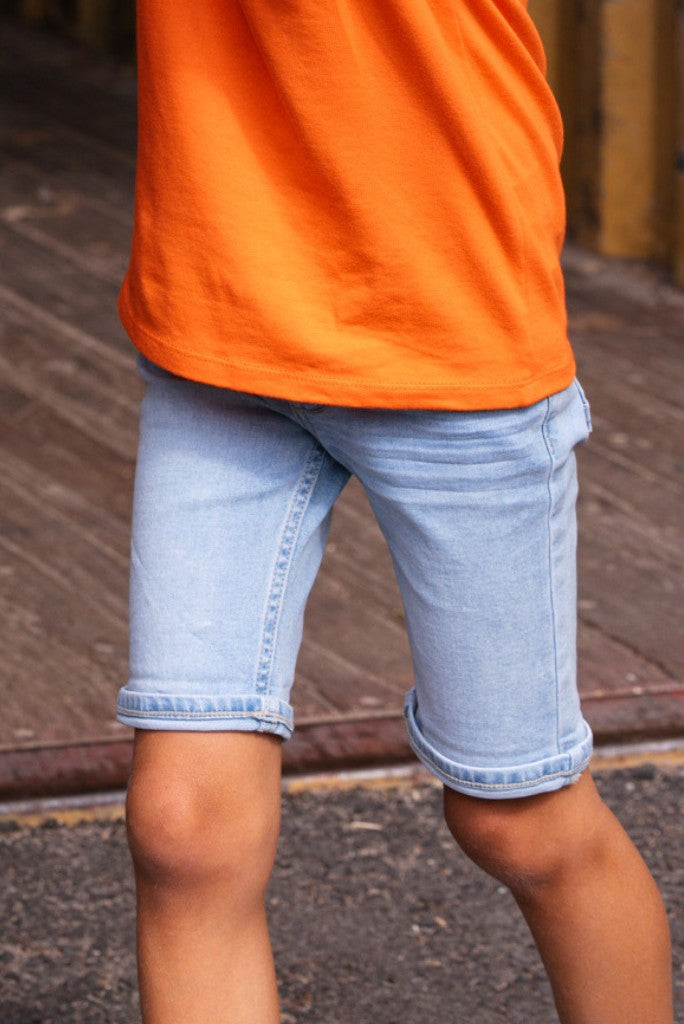 Boy wearing an orange shirt and light blue denim shorts Quinn on a wooden floor.