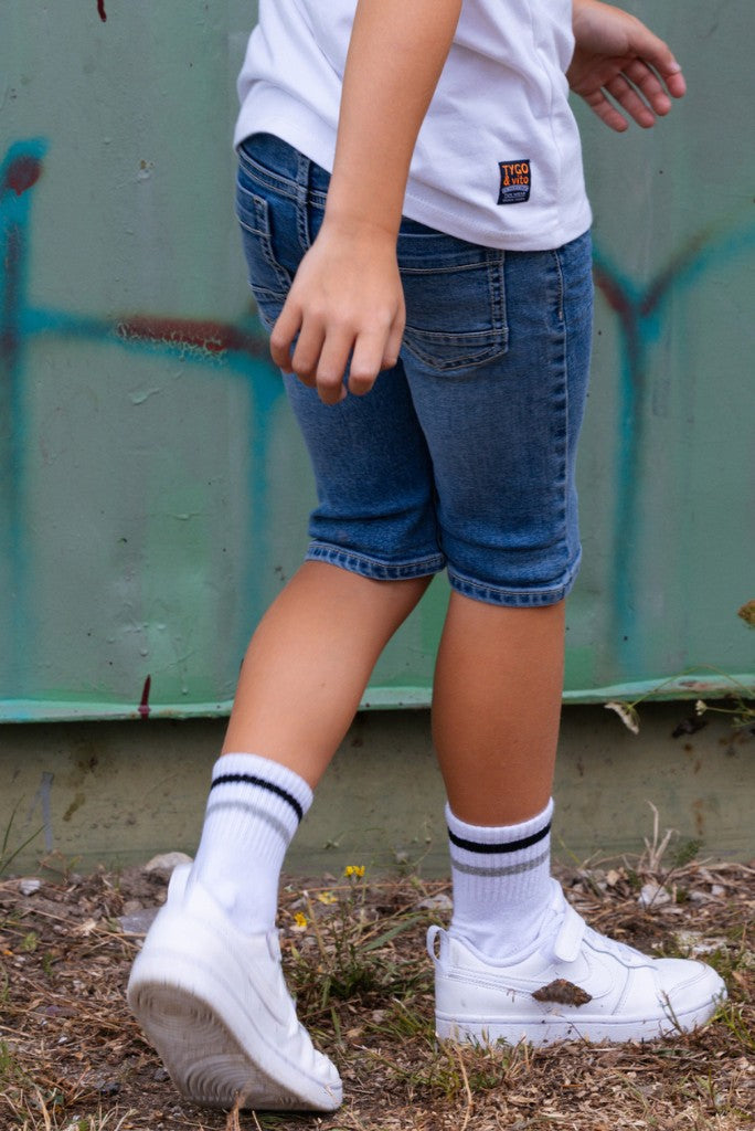 Boy wearing blue shorts Quinn, white shirt, and white sneakers with black stripes in front of a green wall.