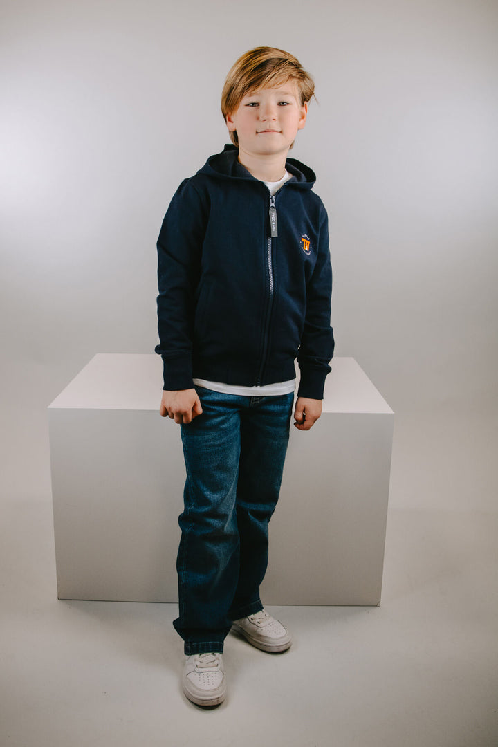 Young boy wearing a navy blue hoodie and jeans standing on a white platform against a plain background