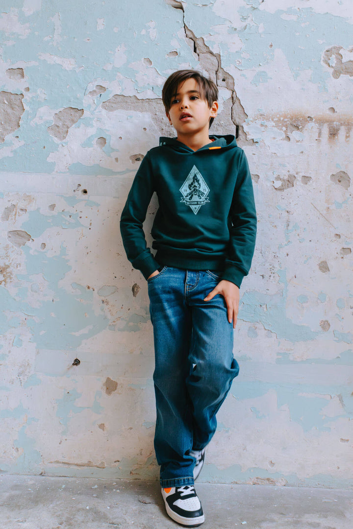 Boy wearing a green hoodie with a logo and blue jeans Loyd against a textured wall.