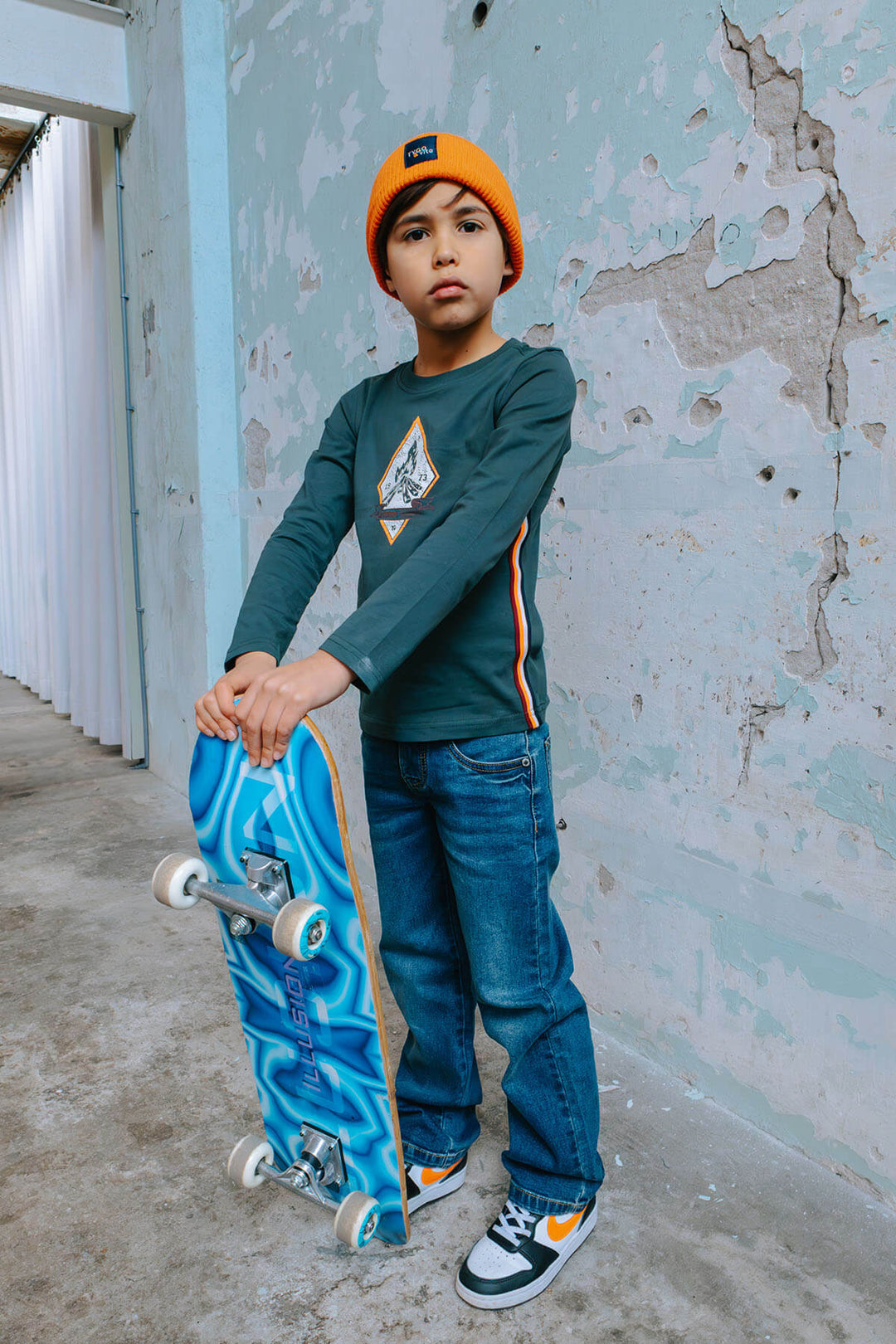 Boy holding a blue skateboard against a textured wall and wearing the blue jeans Loyd.