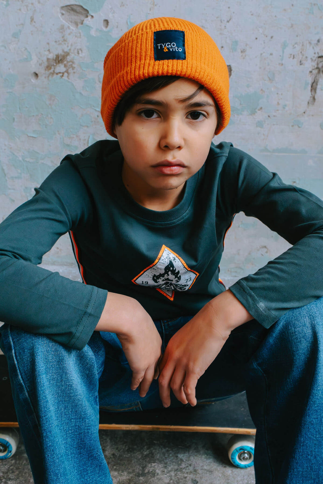 Boy wearing an orange beanie and green long sleeve top with a patch, sitting on a skateboard.