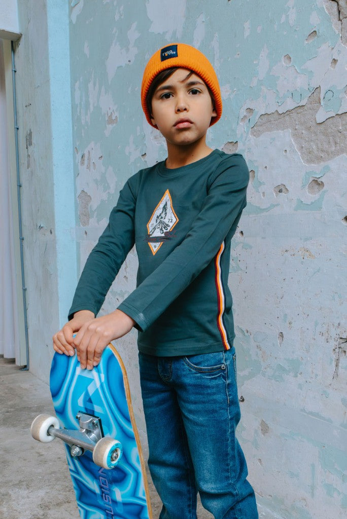 Boy wearing the green long-sleeve top Mason and orange beanie holding a blue skateboard against a textured wall.