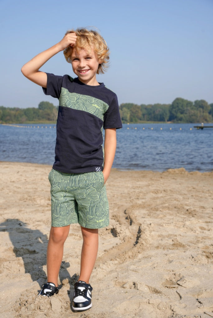 Boy wearing a navy t-shirt Tanner by B.Nosy with green design and patterned shorts standing on a sandy beach.