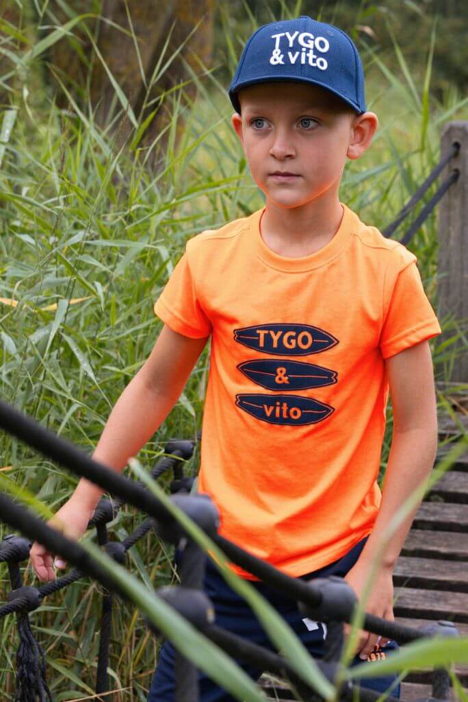 Boy wearing a neon orange T-Shirt James and blue cap with 'Tygo & Vito' branding outdoors.