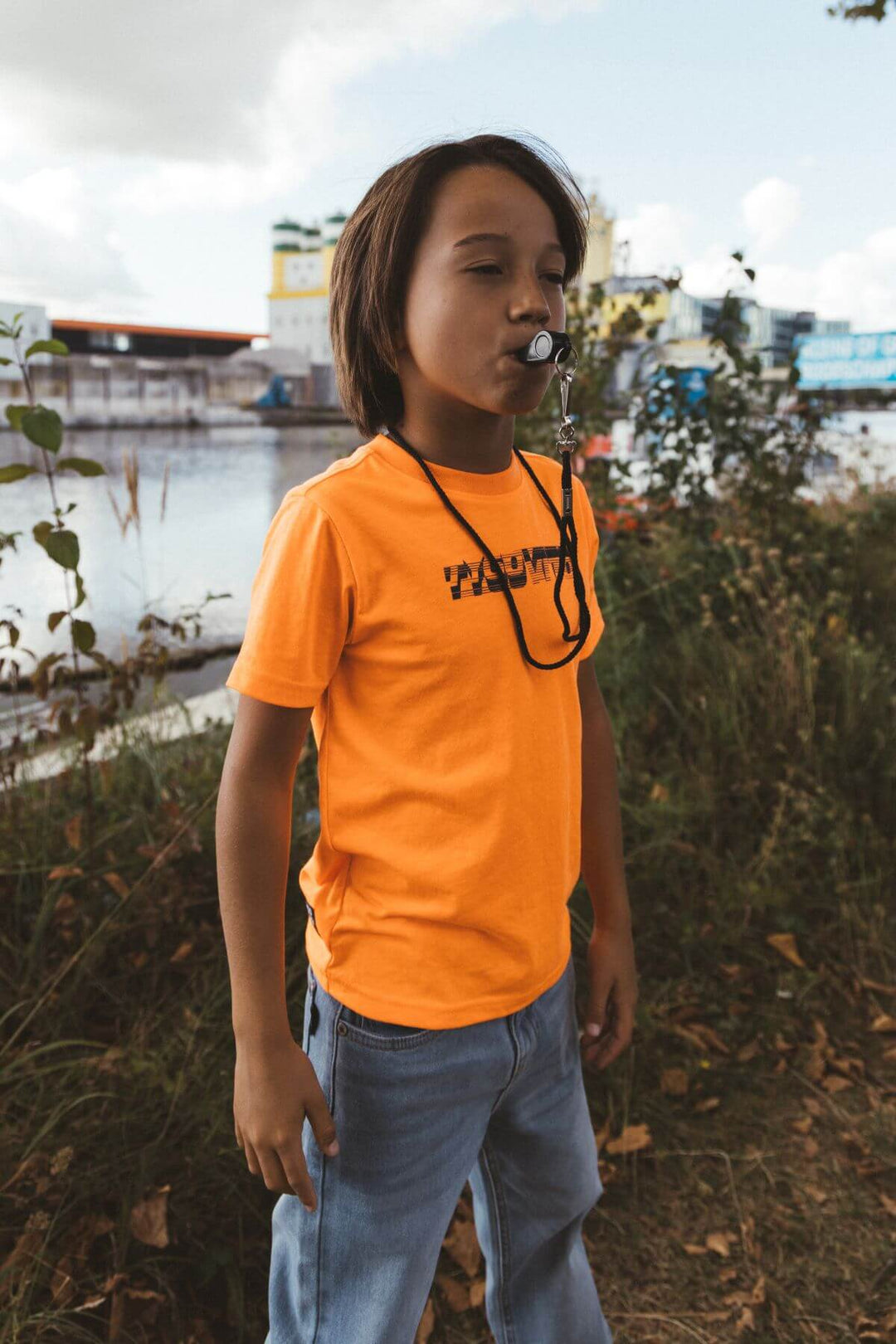 Boy wearing a neon orange T-Shirt with text, standing outdoors by a body of water.