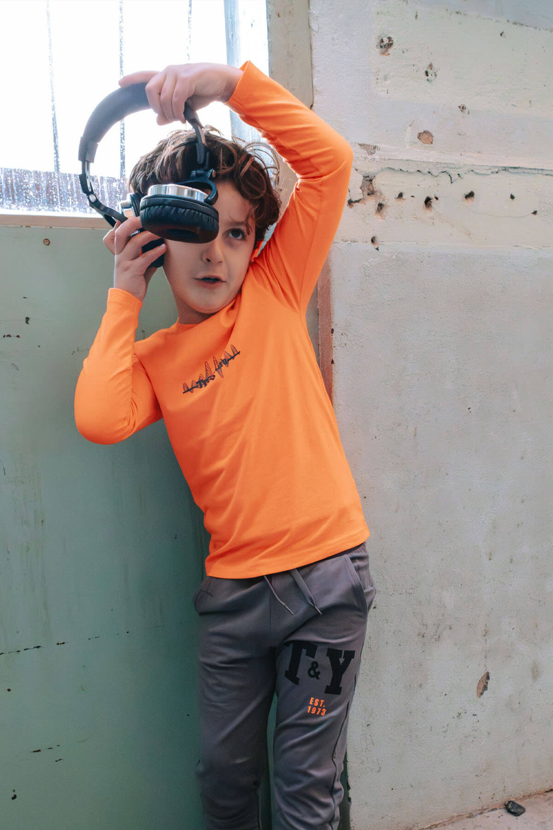 Boy wearing a neon orange shirt and gray pants holding a large black object against a wall.