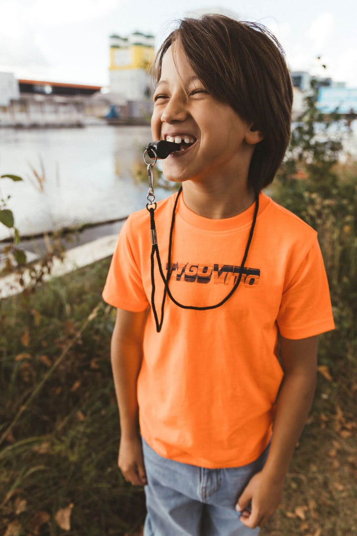 Boy wearing a neon T-Shirt in orange with a lanyard, standing outdoors near a body of water.