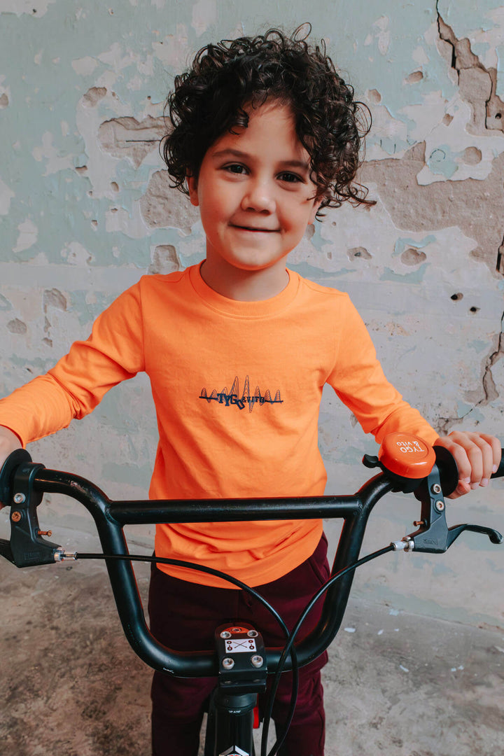 Boy in a neon orange top standing next to a bicycle against a textured wall.