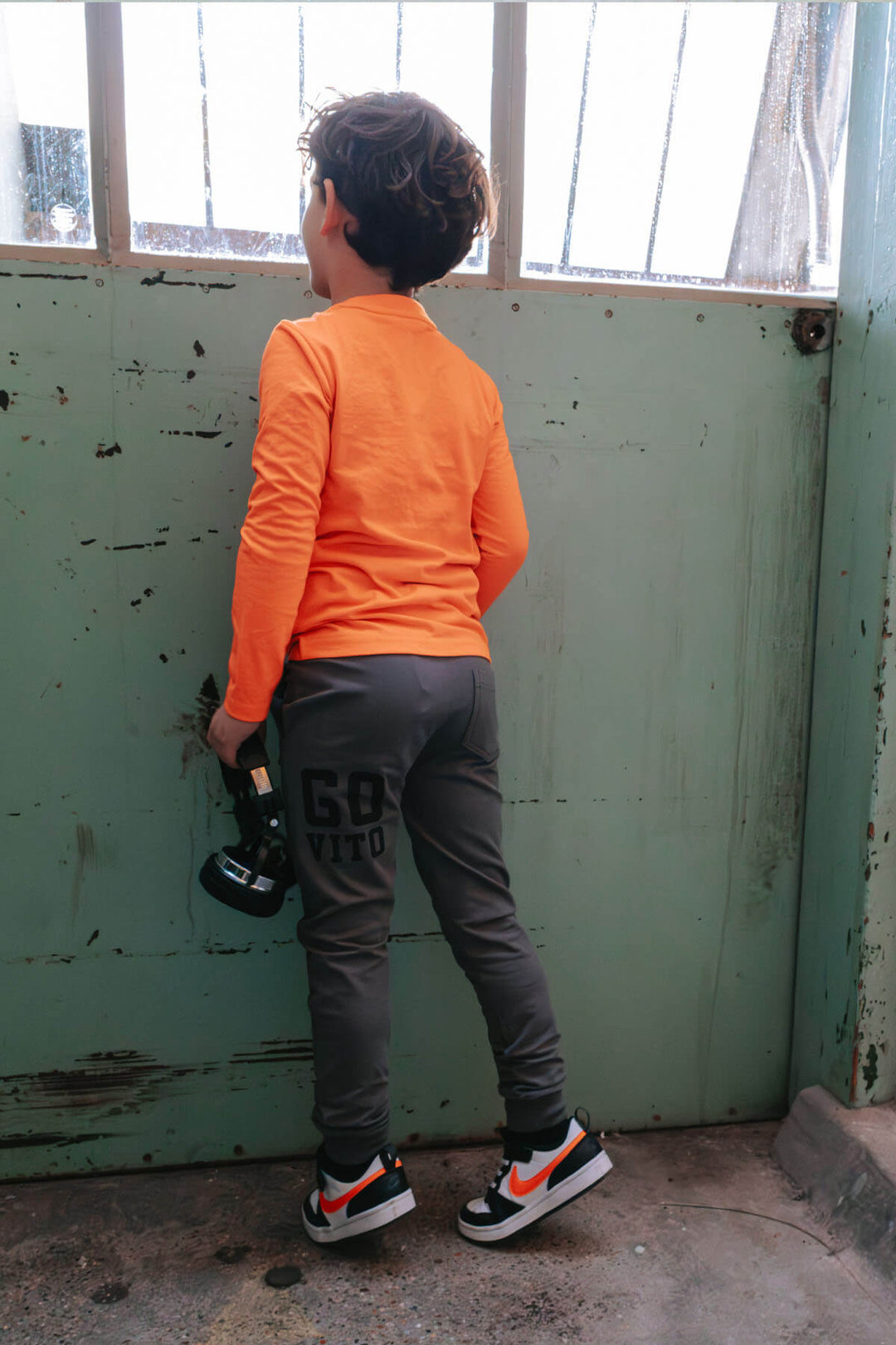 Boy wearing an orange top and gray pants standing in front of a green door.