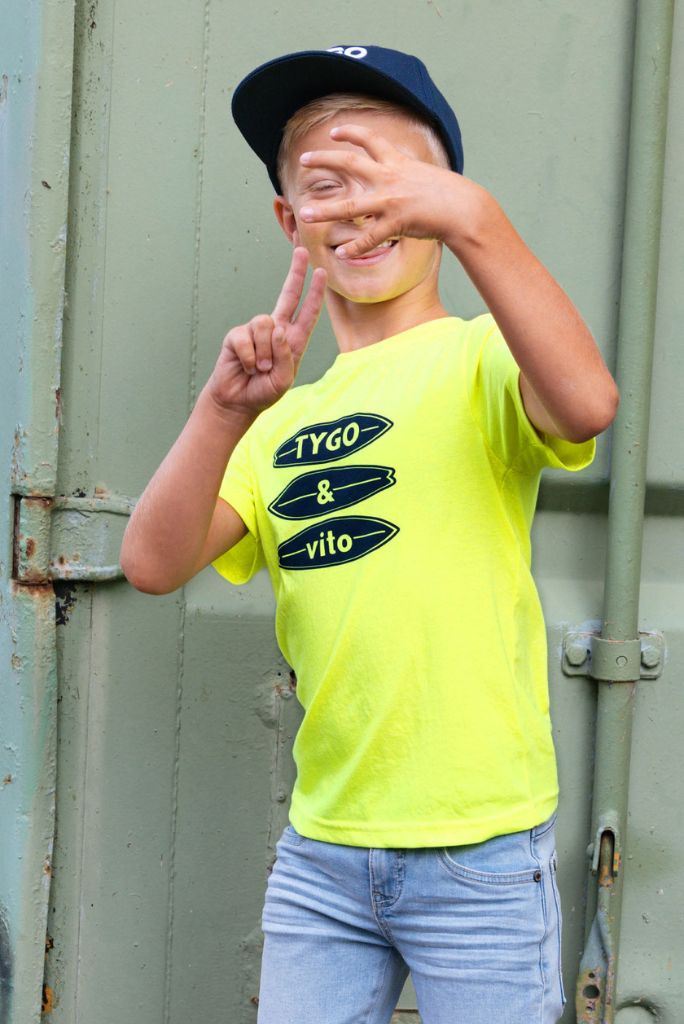 Boy wearing a bright neon  yellow t-shirt James with branding, standing against a metallic background