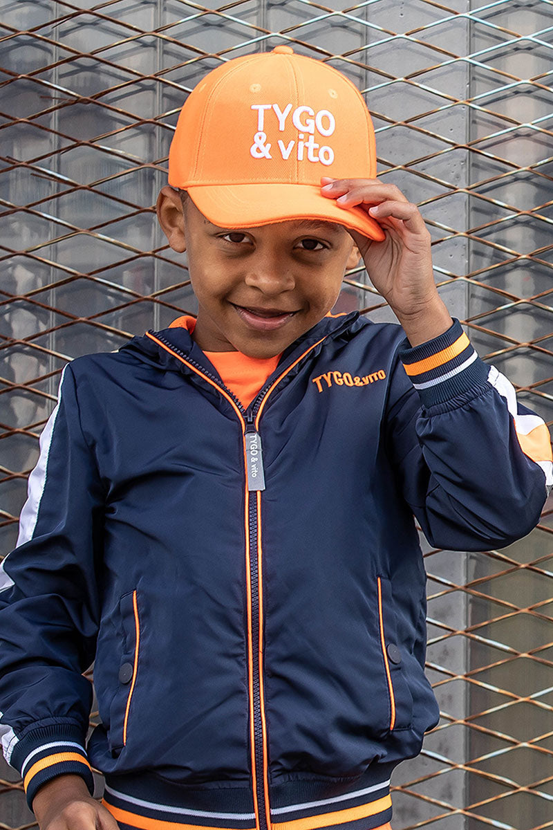 A young boy wearing a navy blue zip-up jacket with orange and white accents, an orange t-shirt, and an orange baseball cap with &