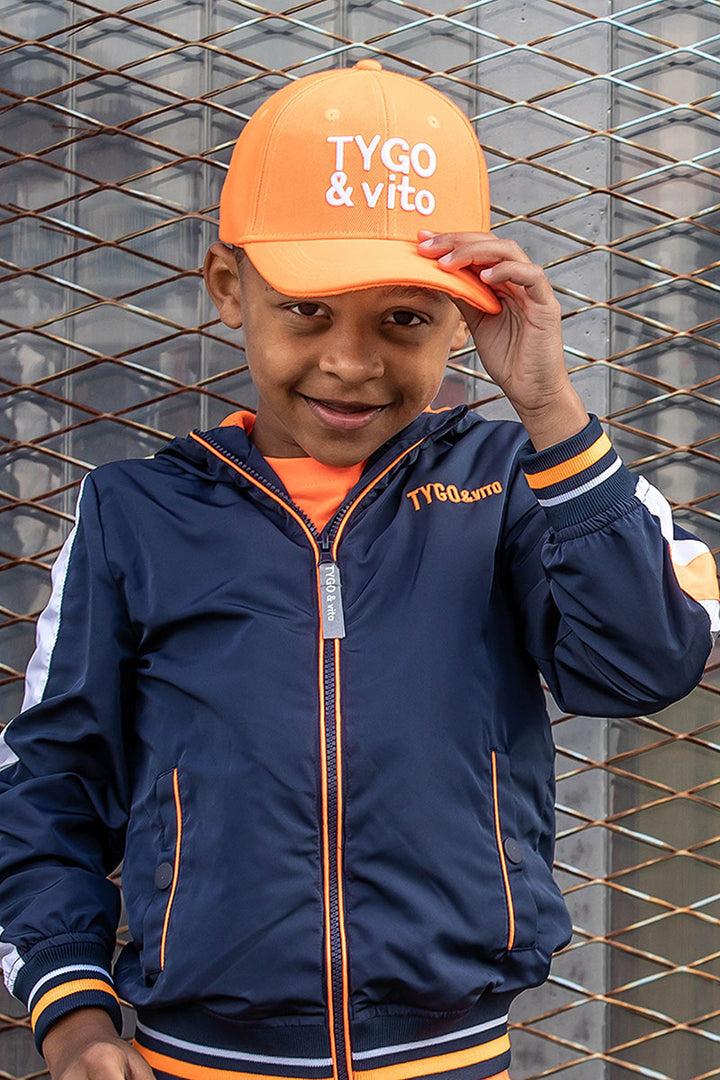 A young boy wearing a navy blue zip-up jacket with orange and white accents, an orange t-shirt, and an orange baseball cap with &