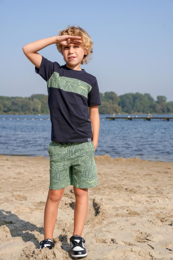 Young boy standing on a beach wearing a black t-shirt with green graphics and patterned shorts Paul by B.Nosy.