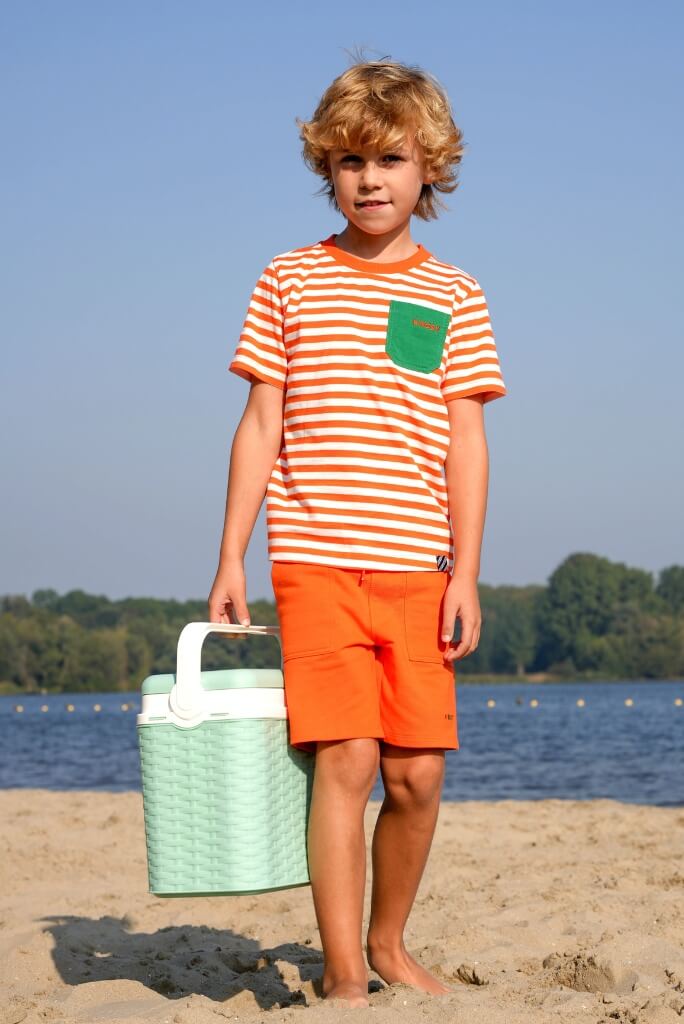 Boy in striped shirt and orange shorts standing on a beach with a cooler.