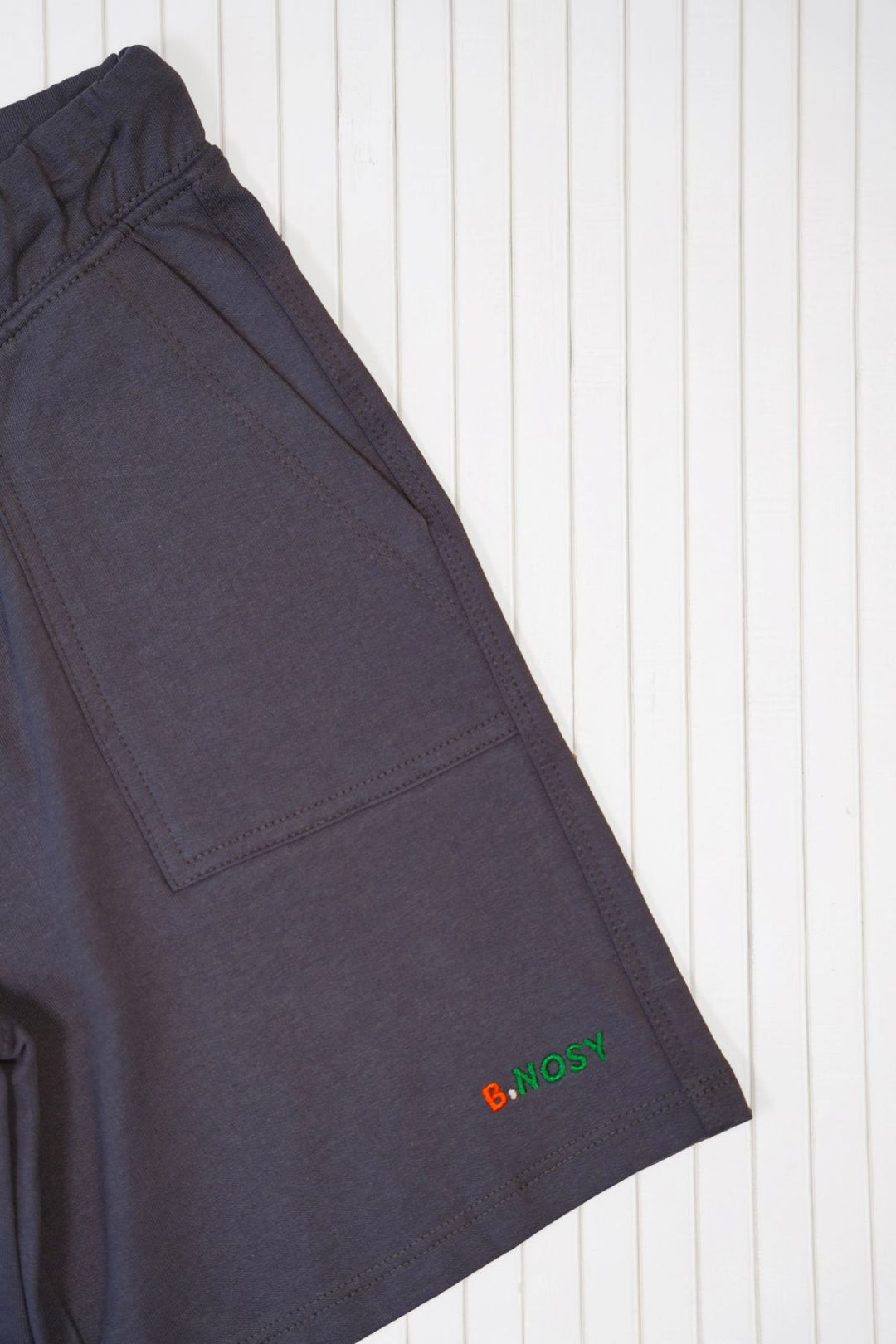 Close-up of dark black shorts laid flat on a white wood plank background, showing a side pocket and a small multicolor embroidered logo near the hem.