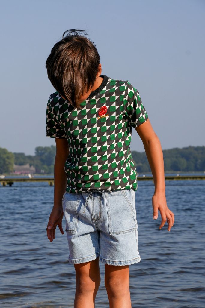 A child standing outdoors by the water wearing a green and black geometric patterned t-shirt with a small red chest graphic and light blue denim shorts.