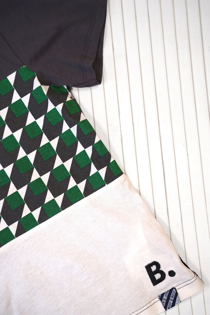 A close-up of a raglan-style t-shirt laid flat on a white slatted surface. The shirt features a dark charcoal gray sleeve, a geometric patterned section with green, black, and white diamonds, and a light beige body with a black embroidered letter &