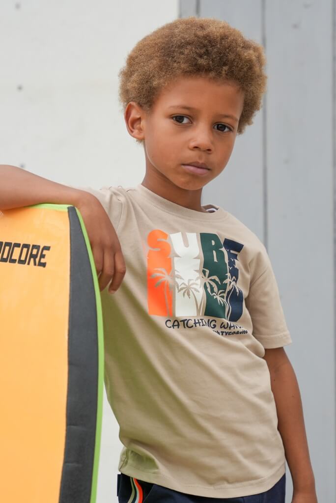 A boy wearing a beige t-shirt Jamiy with graphics, holding a yellow board against a light gray background