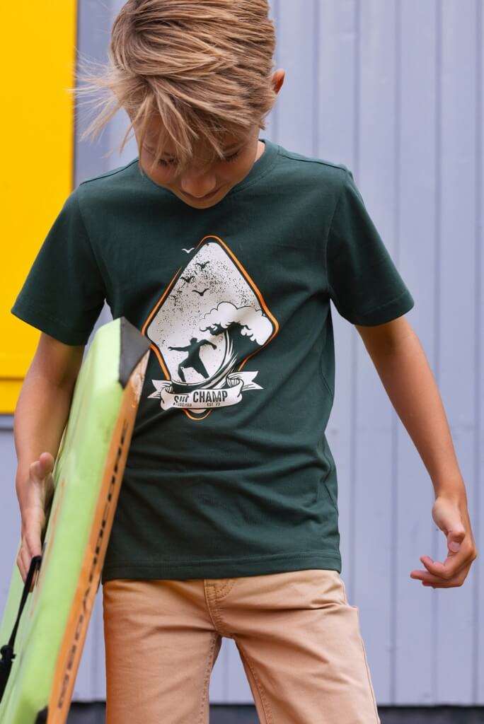 A boy wearing a green t-shirt Jaimy with a graphic design, holding a skateboard against a yellow and gray background.