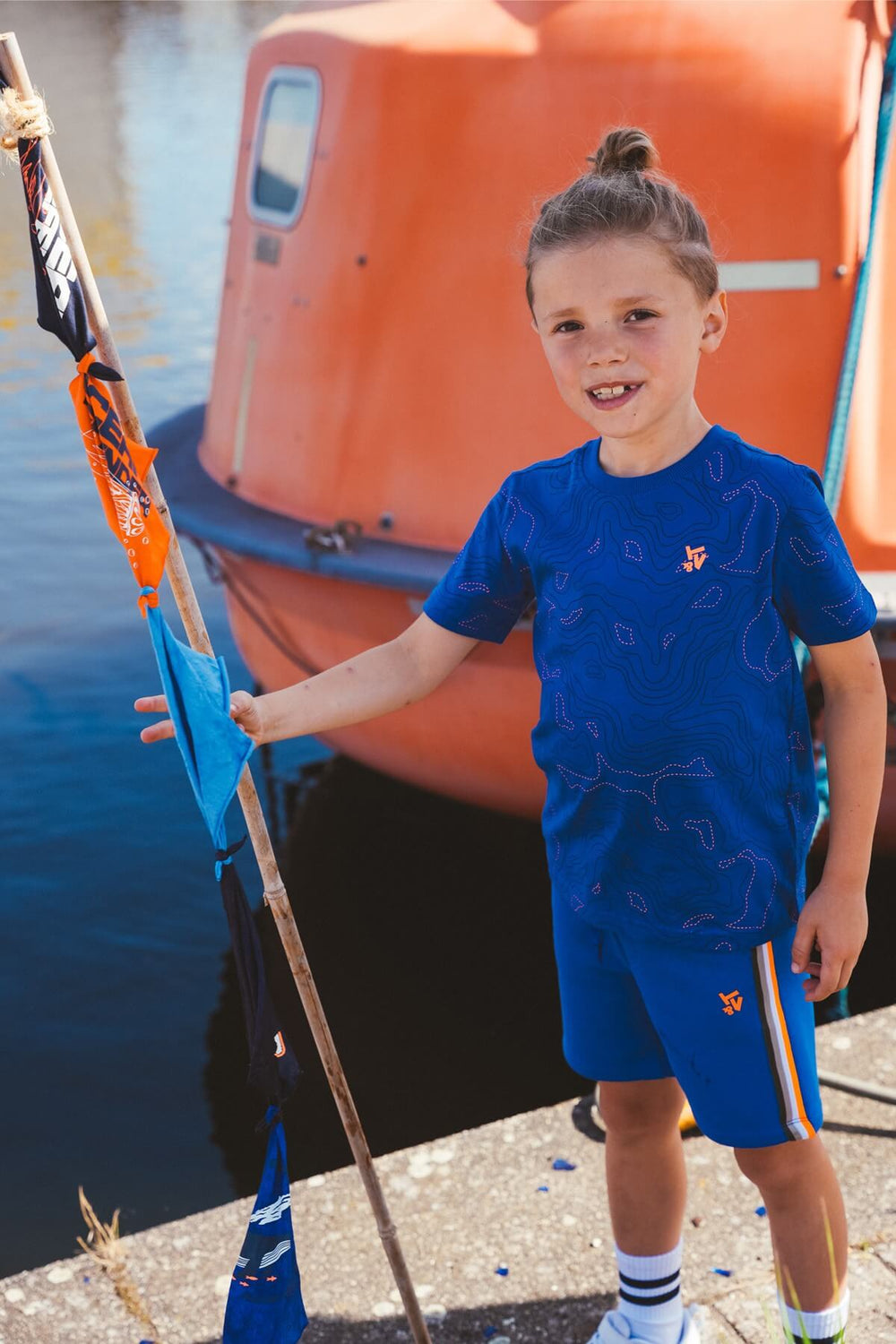 A young boy wearing a blue patterned athletic t-shirt and matching shorts stands on a dock next to water, holding a flag in front of an orange boat.