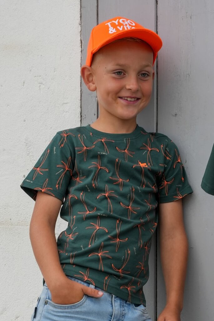 Boy wearing an orange cap and green t-shirt Thijs with red patterns against a gray wooden background