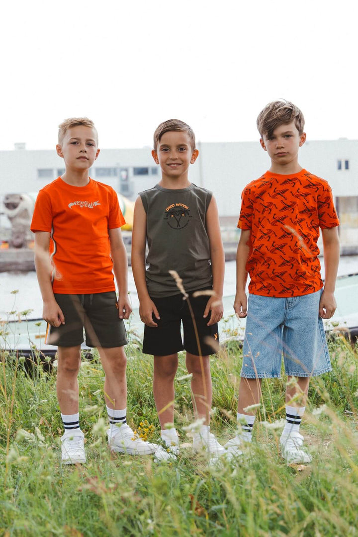 Three boys wearing orange and green shirts standing in a grassy area.
