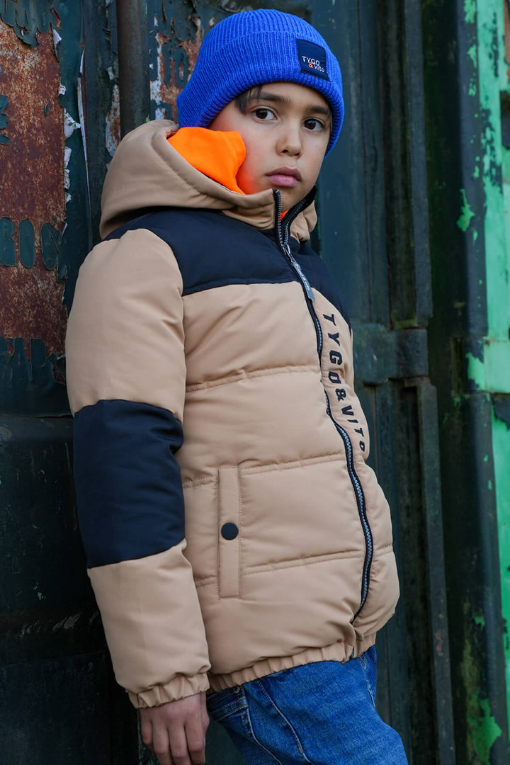 Child wearing a light brown puffer jacket with a blue beanie, standing against a textured wall.