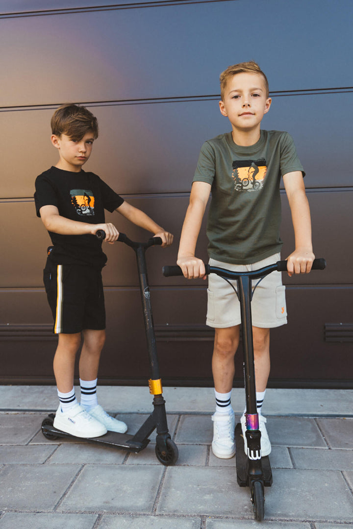 Two boys standing with scooters against a dark wall.
