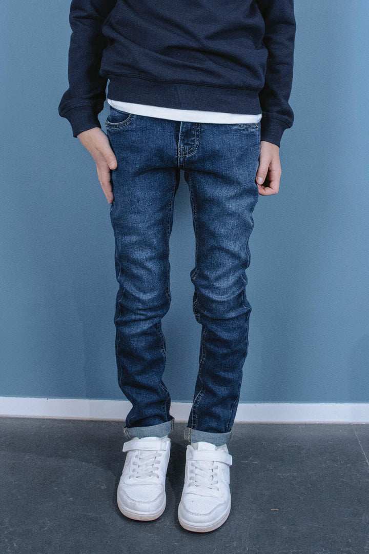 A boy wearing blue skinny jeans and white sneakers against a blue wall.