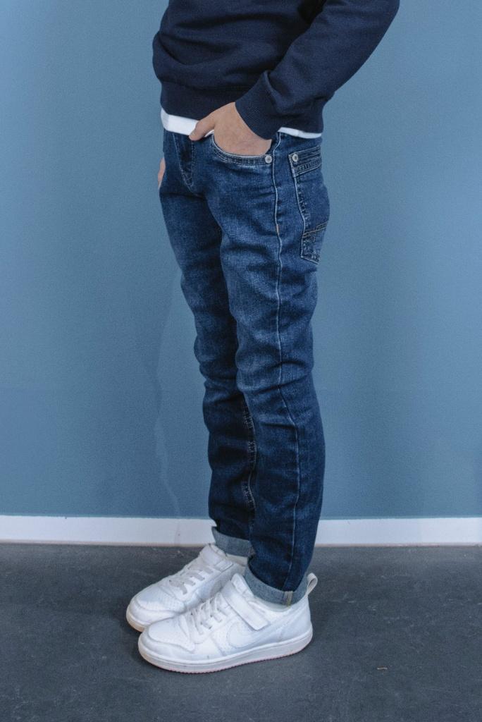 Boy  wearing blue jeans and white sneakers against a blue wall.