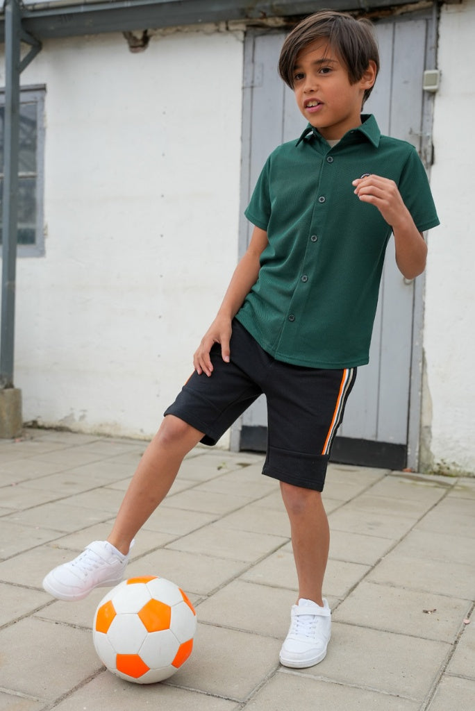 Boy in green shirt and black shorts playing with a soccer ball on a paved surface.