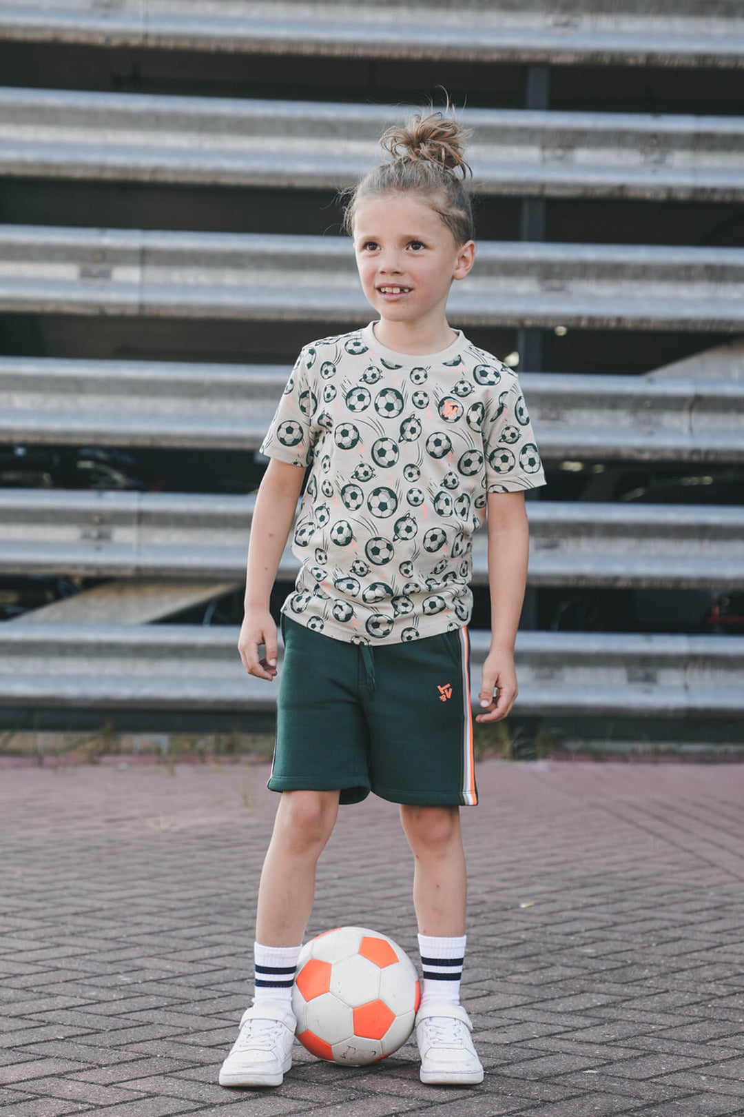 Boy wearing a patterned shirt and green shorts, holding a soccer ball on a paved surface.