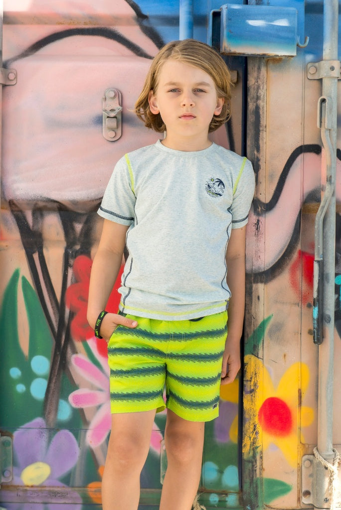 Boy wearing a gray T-shirt Tony by B.Nosy and colorful shorts standing in front of a graffiti-covered wall.