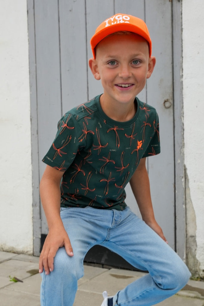 Boy wearing an orange cap and green shirt with red patterns, sitting against a wooden wall.