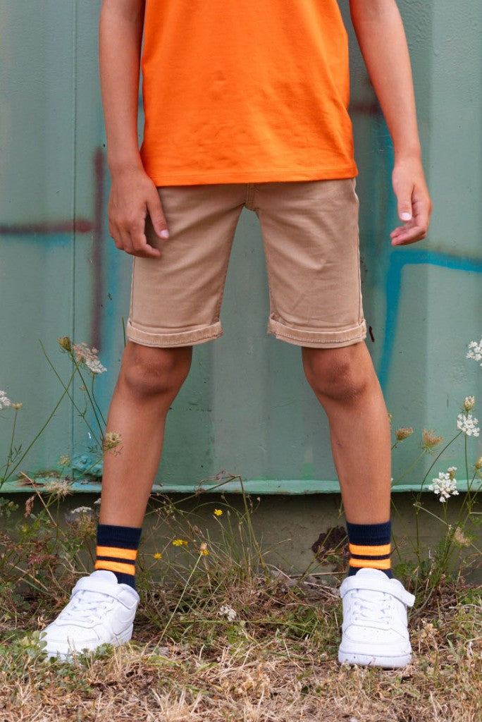 Boy wearing an orange shirt, beige shorts, and white sneakers with black and yellow socks standing on grass.