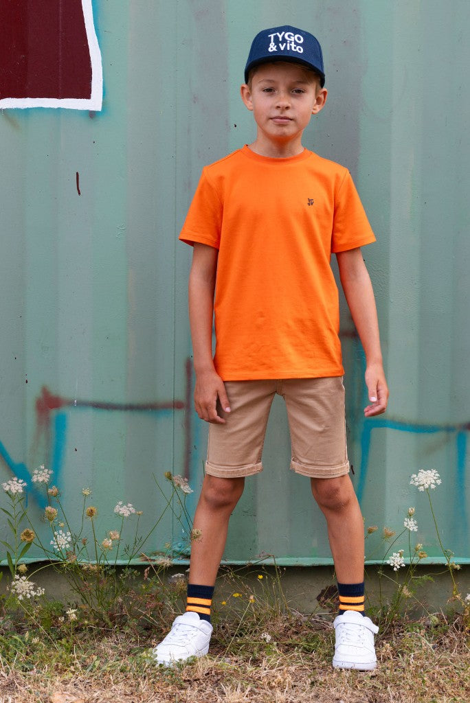 Boy wearing an orange t-shirt and beige shorts Quinn standing in front of a green metal wall.