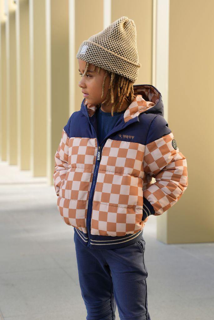 Boy wearing a checkered jacket and beanie standing against a beige wall.
