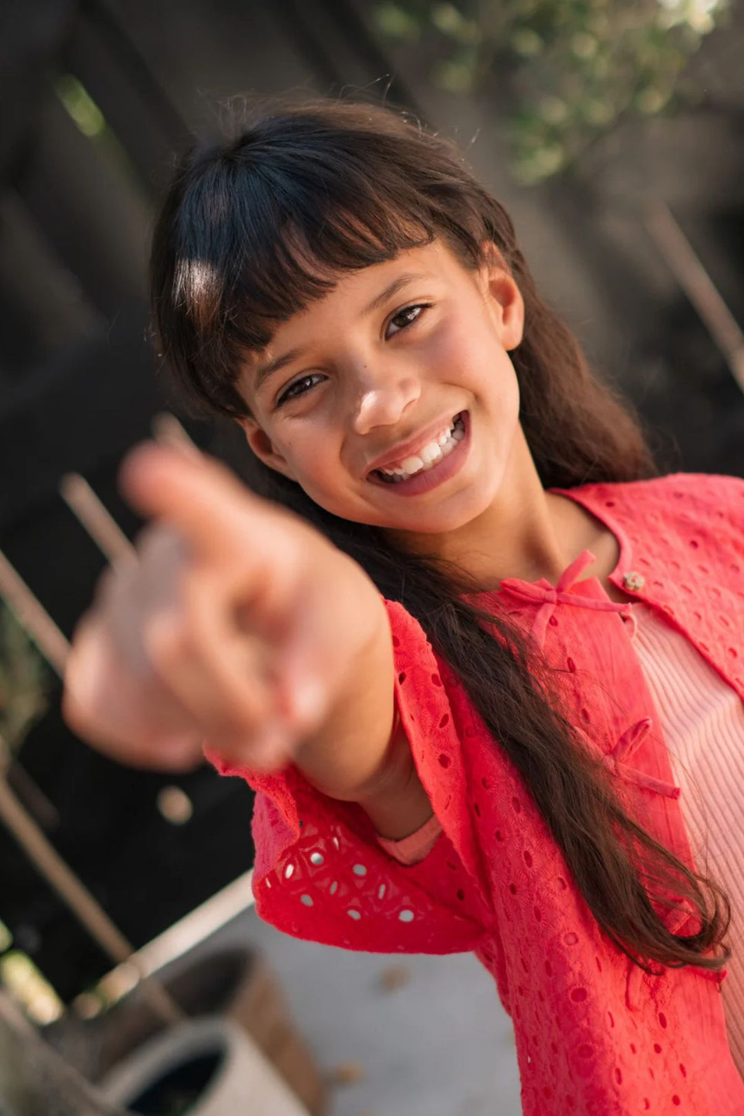 A smiling young girl with dark hair and bangs points directly at the camera, wearing a bright coral pink blouse over a light pink ribbed top. The background is a softly blurred outdoor setting with a wooden fence.