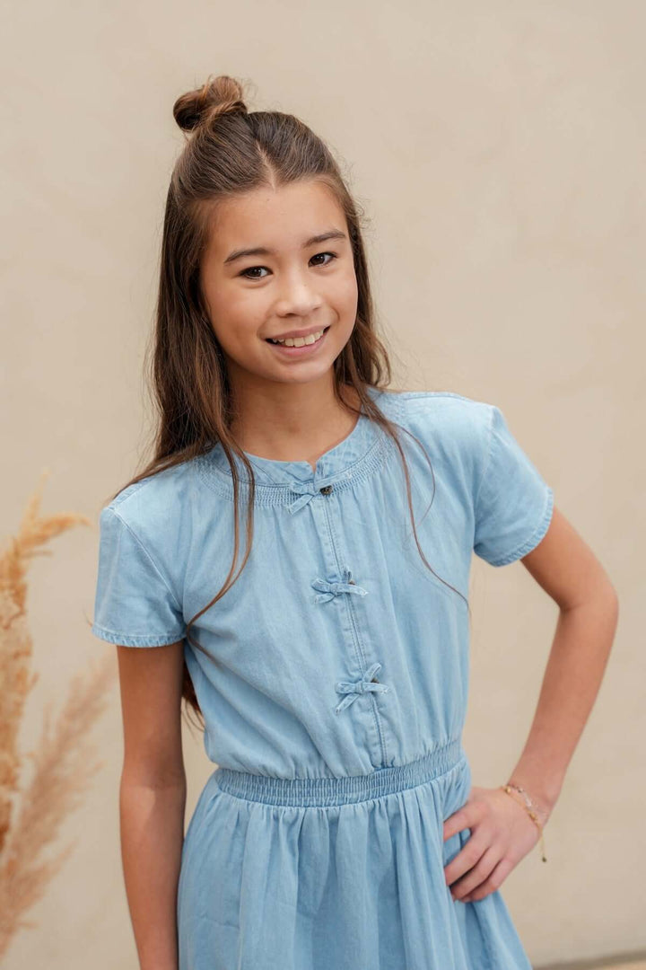 A young girl with her hair in a top bun wearing a light blue short-sleeve dress with button details on the chest, standing against a neutral beige wall.