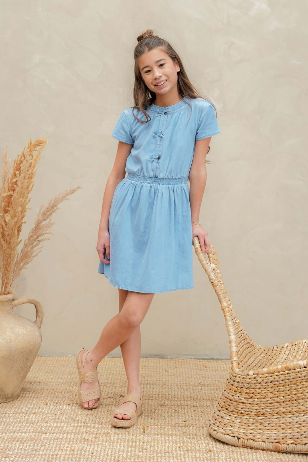 A young girl wearing a light blue short-sleeve button-front dress stands in a styled studio setting with a beige textured wall, woven chair, dried grass in a vase, and a woven rug.