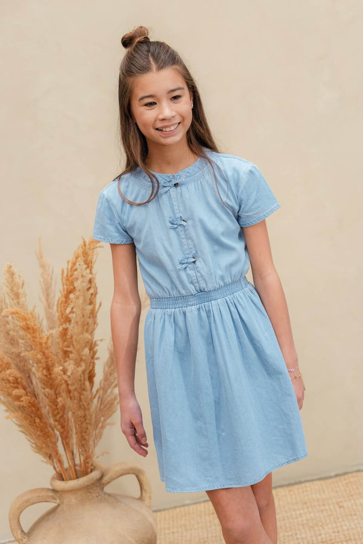 A young girl with her hair in a bun models a light blue short-sleeve dress with a smocked waist and button details on the chest.
