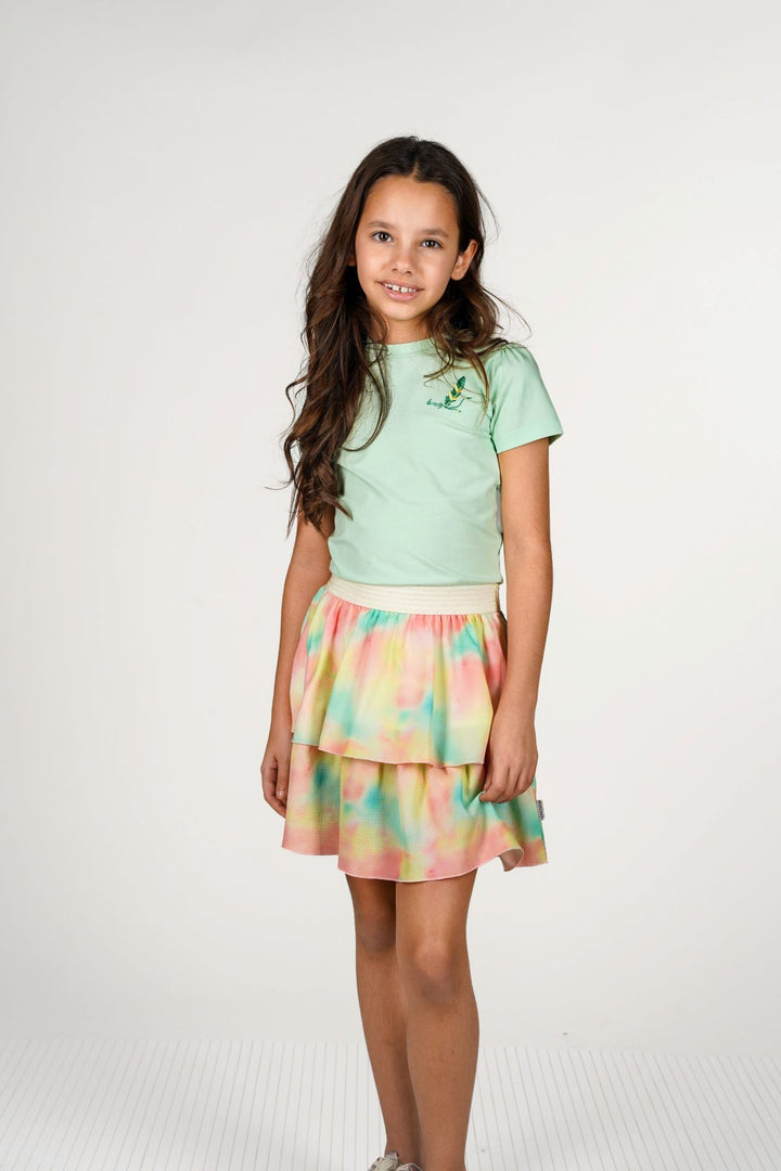 A young girl with long brown hair stands against a light gray studio background, wearing a mint green short-sleeve dress with a tiered pastel tie-dye skirt in pink, yellow, and green.